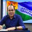 Imagem de um homem calvo com poucos cabelos escuros, ele é branco e usa uma camisa social azul marinho. ele assina um documento sentado em uma mesa. Ao fundo aparece a bandeira de Jaboatão dos Guararapes