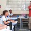 Em uma sala de aula, um professor branco de cabelo castanho escuro aponta para o quadro enquanto fala para vários alunos sentados em cadeiras. Os estudantes usam a farda do Governo de Pernambuco
