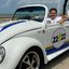 Na praia de Boa Viagem, Fred Ferreira dirige um Fusca moderno na cor branca com detalhes azuis e amarelos. O fusca não tem teto. Fred Ferreira sorri com a mão na lateral do carro