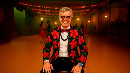 Na foto, o artista renomado Carlinhos de Jesus. Ele está vestido com uma roupa formal e chamativa, característica da dança de salão, sorrindo. Sentado e ao fundo um salão com pessoas dançando.