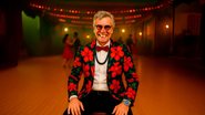 Na foto, o artista renomado Carlinhos de Jesus. Ele está vestido com uma roupa formal e chamativa, característica da dança de salão, sorrindo. Sentado e ao fundo um salão com pessoas dançando.