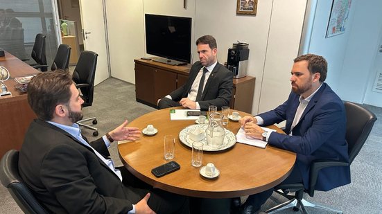 Eduardo Moura e Técio Teles sentados em um mesa em frente a um homem branco de barba, parece um escritório
