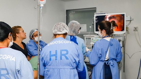 Diversos profissionais com aventais de hospiatis, pessoas usam máscaras e toucas. Avental diz endoscopia HR