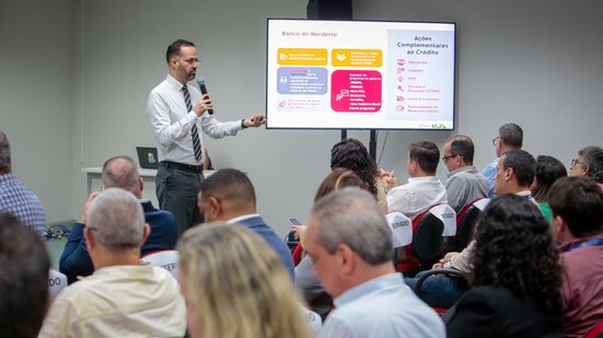 Diversas pessoas sentadas olham para Hugo Queiroz, que fala com um microfone ao lado de uma televisão com nome do banco do nordeste e números fora de foco