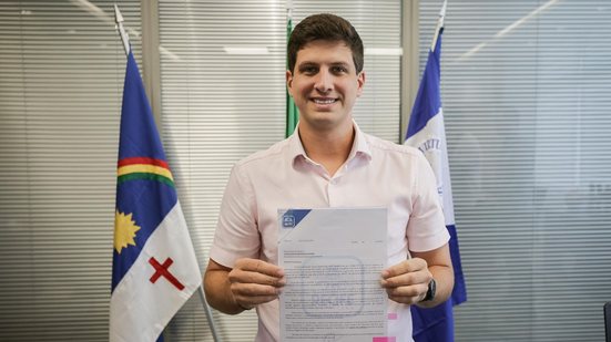 João Campos sorri enquanto segura um documento. Ao fundo as bandeiras de Pernambuco e do Recife