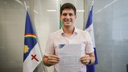 João Campos sorri enquanto segura um documento. Ao fundo as bandeiras de Pernambuco e do Recife