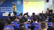 Homem em um palco falando em um microfone e plateia de alunos