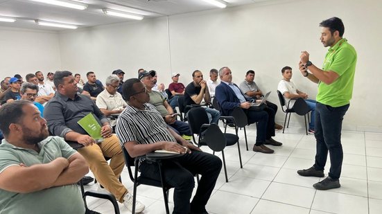 Homem com camiseta verde em pé fala com diversas pessoas sentadas prestando atenção nele. Sala de aula