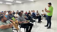 Homem com camiseta verde em pé fala com diversas pessoas sentadas prestando atenção nele. Sala de aula