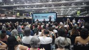 Palco com diversas pessoas sentadas. Símbolo da Bienal escrito ViaOmar