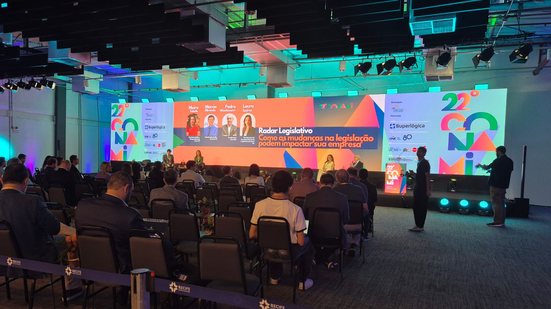 Telão escrito ""Radar Legislativo: Como as mudanças na legislação podem impactar sua empresa" e fotos dos palestrantes, em frente pessoas sentadas observando