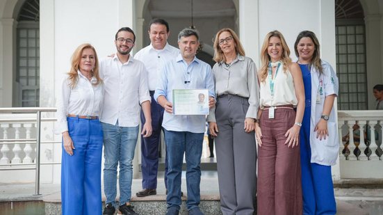 Segurando certificado com seu rosto, Eduardo da Fonte sorri ao lado de Lula da Fonte, 3 mulheres loiras e um homem, eles estão em frente a uma escada do imip