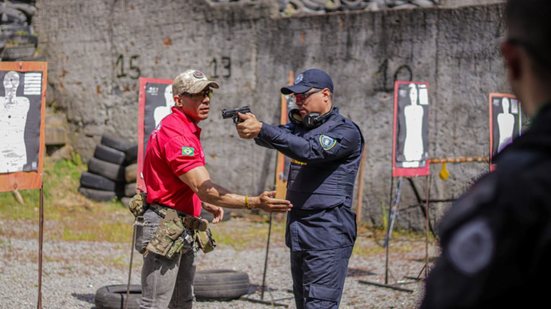 Homem com farda da guarda municipal segura arma enquanto outro ajuda a posicioná-lo, ao fundo alvos de papel