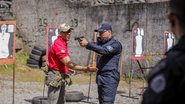 Homem com farda da guarda municipal segura arma enquanto outro ajuda a posicioná-lo, ao fundo alvos de papel