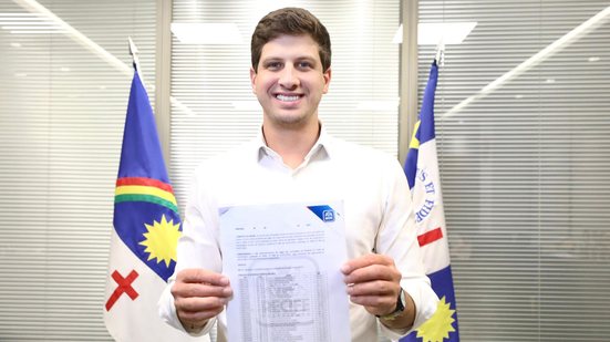 João Campos sorri com documento em mãos. Ao fundo bandeira do Recife e de Pernambuco