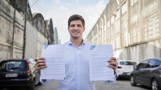 João Campos segura dois documentos, um em cada mão, enquanto sorri. Ao fundo áreas mais degradadas