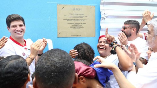 João Campos bate palmas em frente a placa de praça, diversas pessoas sorriem ao redor, ao fundo Humberto Costa bate palmas