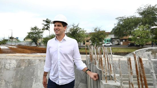 João Campos com capacete de construção coloca a mão em lateral de concreto com algumas estacas