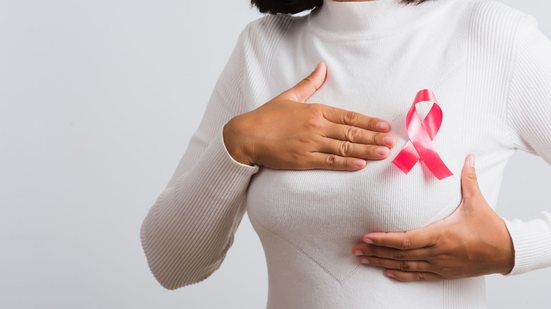Moça segurando o seio (vestida com blusa de manga comprida) com laço do Outubro Rosa entre as mãos