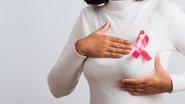 Moça segurando o seio (vestida com blusa de manga comprida) com laço do Outubro Rosa entre as mãos