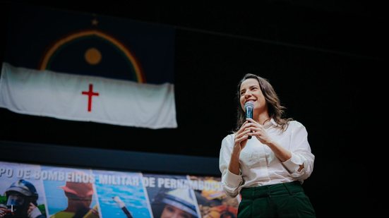 Raquel Lyra falando em microfone com bandeira de Pernambuco ao fundo