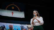 Raquel Lyra falando em microfone com bandeira de Pernambuco ao fundo