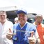 Ricardo Paes Barreto sorri e faz "joinha" com duas mãos, ele usa colete e boné azuis. Homens com blusas brancas estão ao seu lado, o sol é forte