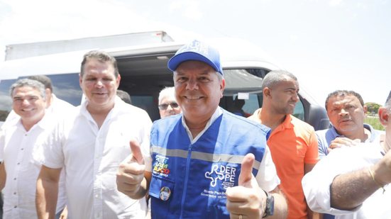 Ricardo Paes Barreto sorri e faz "joinha" com duas mãos, ele usa colete e boné azuis. Homens com blusas brancas estão ao seu lado, o sol é forte