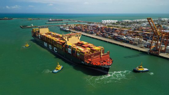 Visão de cima de área de mar com embarcação, na lateral diversas estruturas mecânicas em terra