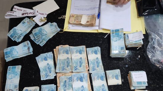 Notas de 100 e 50 reais em uma mesa, ao fundo logo do mppe