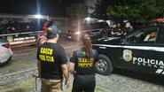 Um homem e uma mulher em pé de costas. Farda do homem está escrito "Gaeco polícia", da mulher está escrito "Gaeco MPPE". Ao fundo viatura da polícia militar. Está escuro
