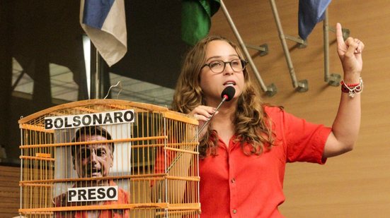 Kari Santos fala em microfone de plenário ao lado de gaiola com a foto de Bolsonaro e escrito "Bolsonaro preso"