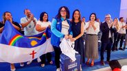 Raquel Lyra sorri ao lado de professor com colete do ganhe o mundo, ao fundo pessoas com a bandeira de pernambuco sorriem e batem palmas