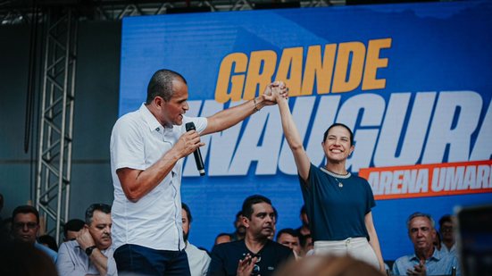 Raquel Lyra sorri em pé e está com as mãos atadas com Janjão, que fala ao microfone. Ao fundo pessoas batem palmas, no telão há "grande inauguração"