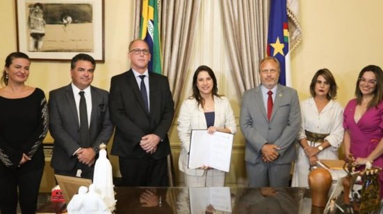 Raquel Lyra, Renato Márcio Rocha Leite, Tulio Vilaça, um homem de terno e três mulheres sorriem em pé lado a lado. Raquel segura um documento