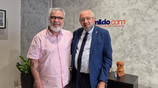 Jamildo e Almir Cirilo em pé sorrindo abraçados, ao fundo sala com parede cinza com logo do Jamildo.com em azul e laranja