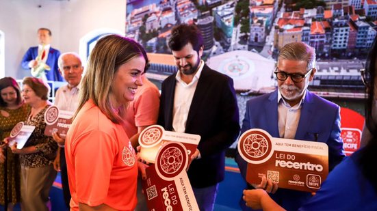 Ana Paula Vilaça sorri enquanto entrega a pessoas placas escrito imóvel isento lei do recentro