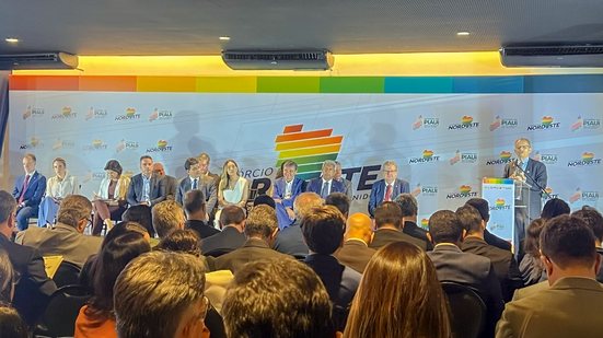 Diversas pessoas sentadas em um painel com fundo consórcio nordeste, em frente pessoas sentadas