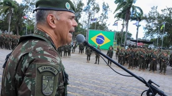 Raquel Lyra ajoelhada sorri enquanto está com as mãos em uma muda de planta recém enterrada, vários homens, alguns com farda do Exército observam
