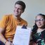 João Campos ao lado de uma mulher na cadeira de rodas, eles sorriem enquanto mostram documento assinado