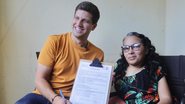 João Campos ao lado de uma mulher na cadeira de rodas, eles sorriem enquanto mostram documento assinado