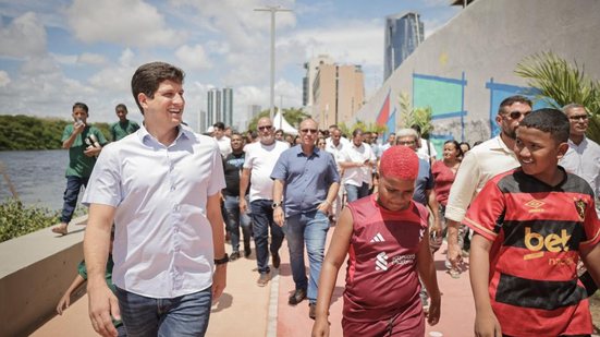 João Campos ao lado de 2 crianças, ao fundo de diversas pessoas andam