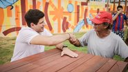 Sentado em mesa de madeira, João Campos cumprimenta um homem de meia idade negro que usa um boné, ao fundo parede pintada colorida em um ambiente externo com grama