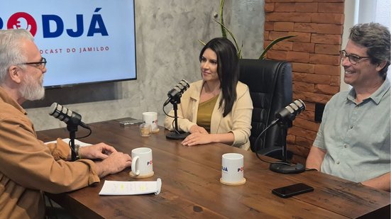Sentados em mesa em frente a microfones, estão Jamildo, Bruna Claudino e Marcelo Garcia, eles conversam. Ao fundo logo do PodJá