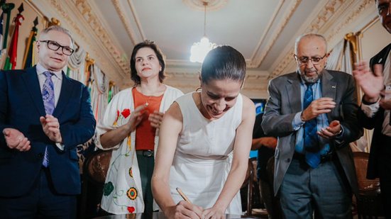 Raquel Lyra assina enquanto Priscila Krause, Almir Cirilo e um homem branco de óculos e careca batem palmas, eles estão em salão do palácio
