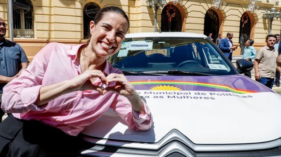 Raquel Lyra sorri enquanto faz coração com a mão. Ela está em frente a carro com bandeira de pernambuco com fundo roxo escrito proteção para mulher