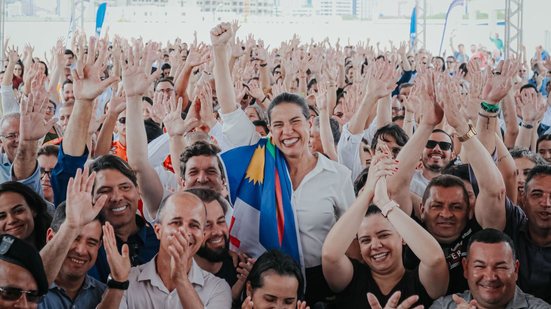 Raquel Lyra levanta braço com sorriso e bandeira de pernambuco no ombro, ela está ao redor de várias pessoas que sorriem e batem palma