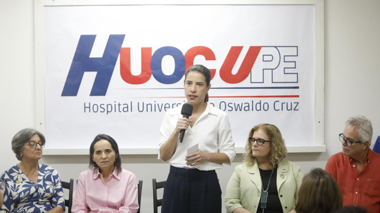 Raquel Lyra em pé fala no microfone ao lado de 3 mulheres e um homem, que estão sentados. Ao fundo simbolo do hospital universitário Oswaldo Cruz e o símbolo da UPE