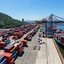 Porto para navios comerciais de grande porte. Na foto há guindastes e contêiner, além de um pico com árvores.