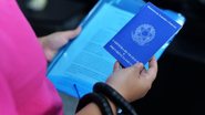 Mulher de costas segurando uma pasta azul com documentos na mão esquerda, à mão direita, ela segura uma Carteira de Trabalho.